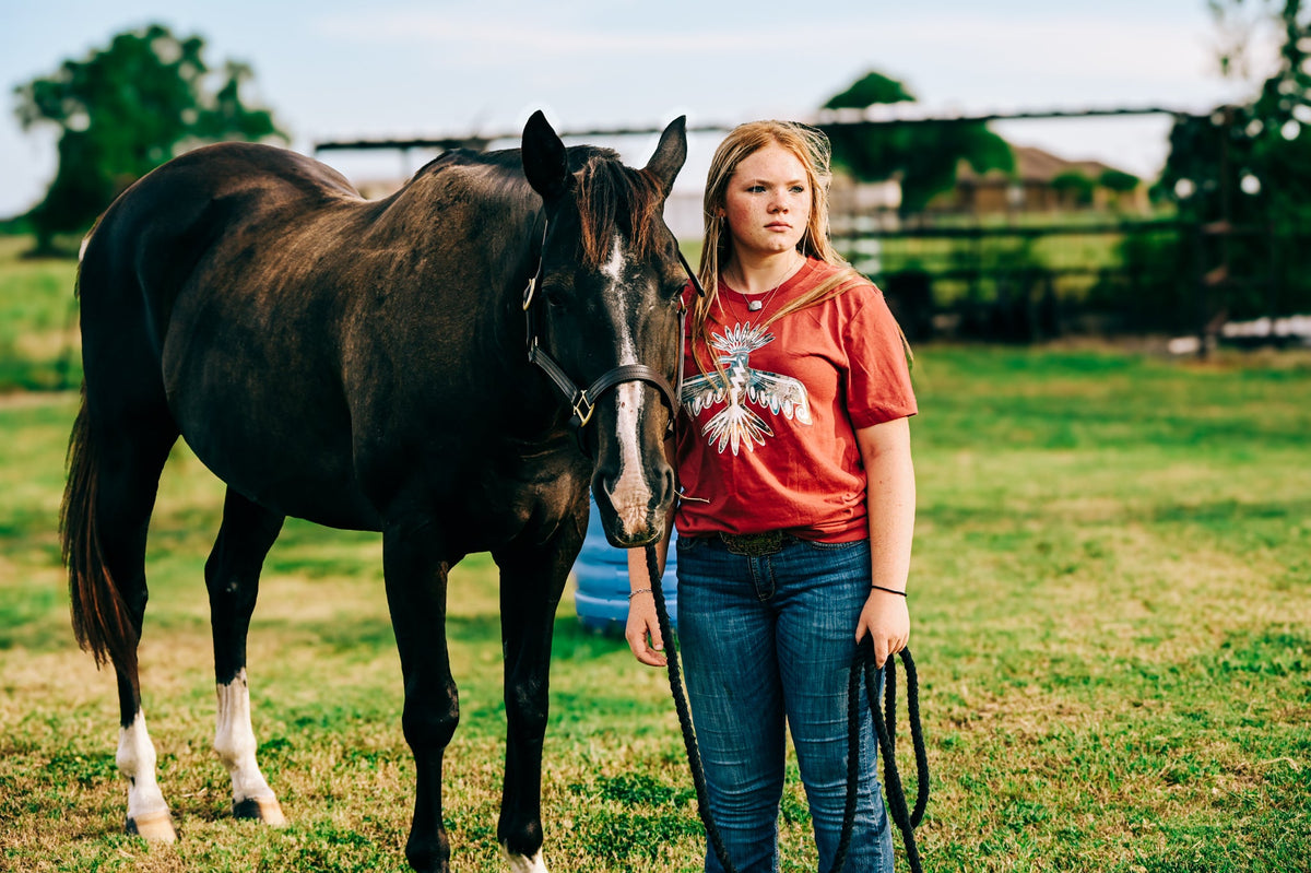 A woman standing in a grassy field next to a dark brown horse, wearing a Mauve Western graphic tee styled with jeans