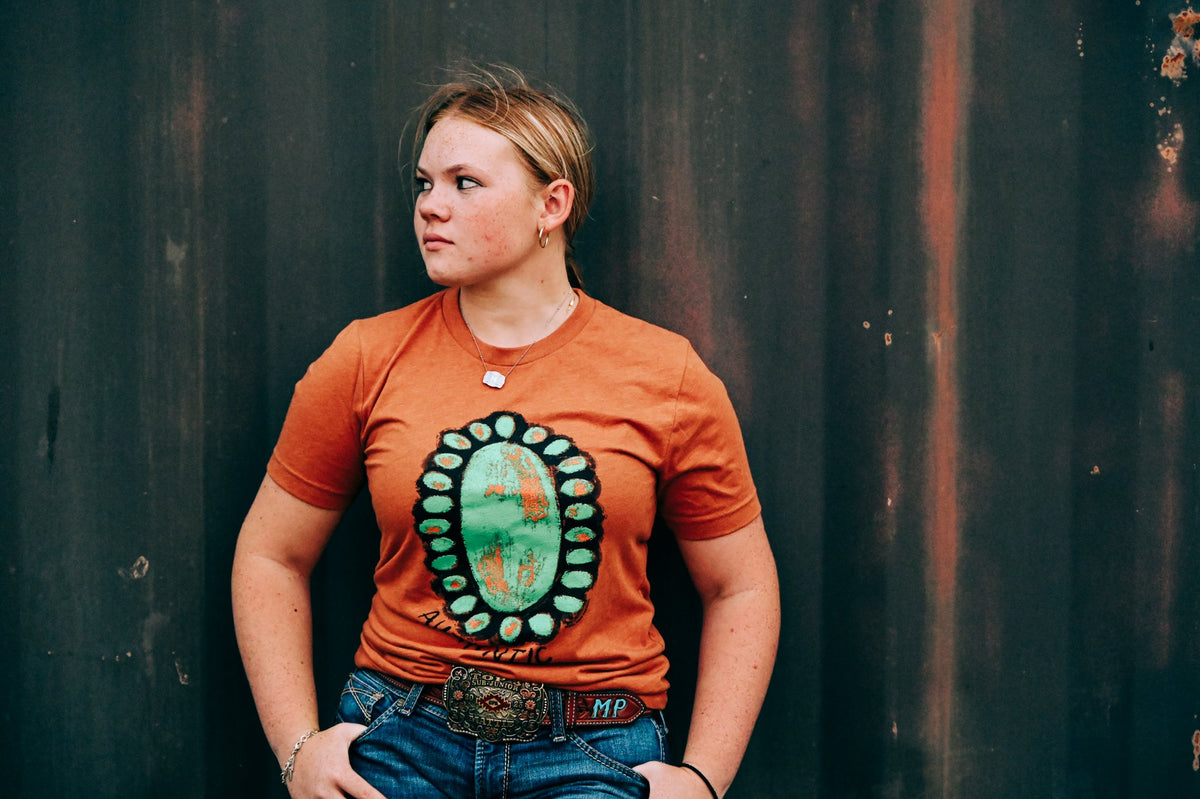 A woman wearing an orange crewneck t-shirt with a large green and orange Southwestern-style geometric graphic on the front, paired with blue jeans and a western belt, standing against a dark corrugated metal wall.