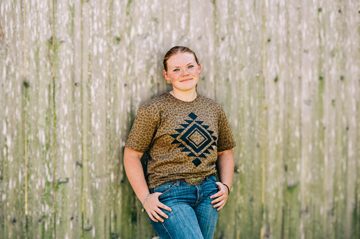 A young girl with blonde hair tied back stands against a weathered wooden fence, wearing a brown and black patterned t-shirt with a geometric diamond design, paired with blue jeans.