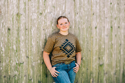 A young girl with blonde hair tied back stands against a weathered wooden fence, wearing a brown and black patterned t-shirt with a geometric diamond design, paired with blue jeans.