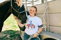 A woman standing next to a horse trailer with a dark horse, wearing a white graphic t-shirt with a cowgirl riding a red horse design and blue jeans.