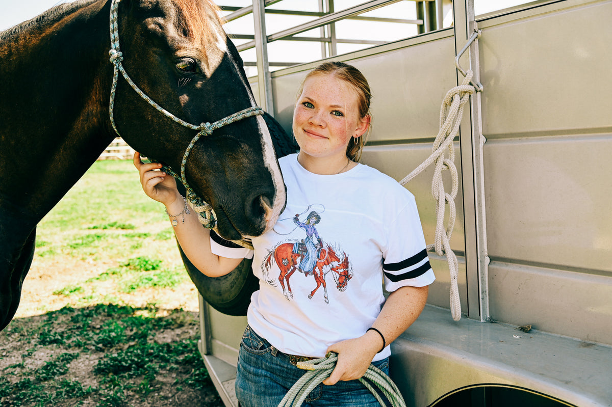 A woman standing next to a horse trailer with a dark horse, wearing a white graphic t-shirt with a cowgirl riding a red horse design and blue jeans.