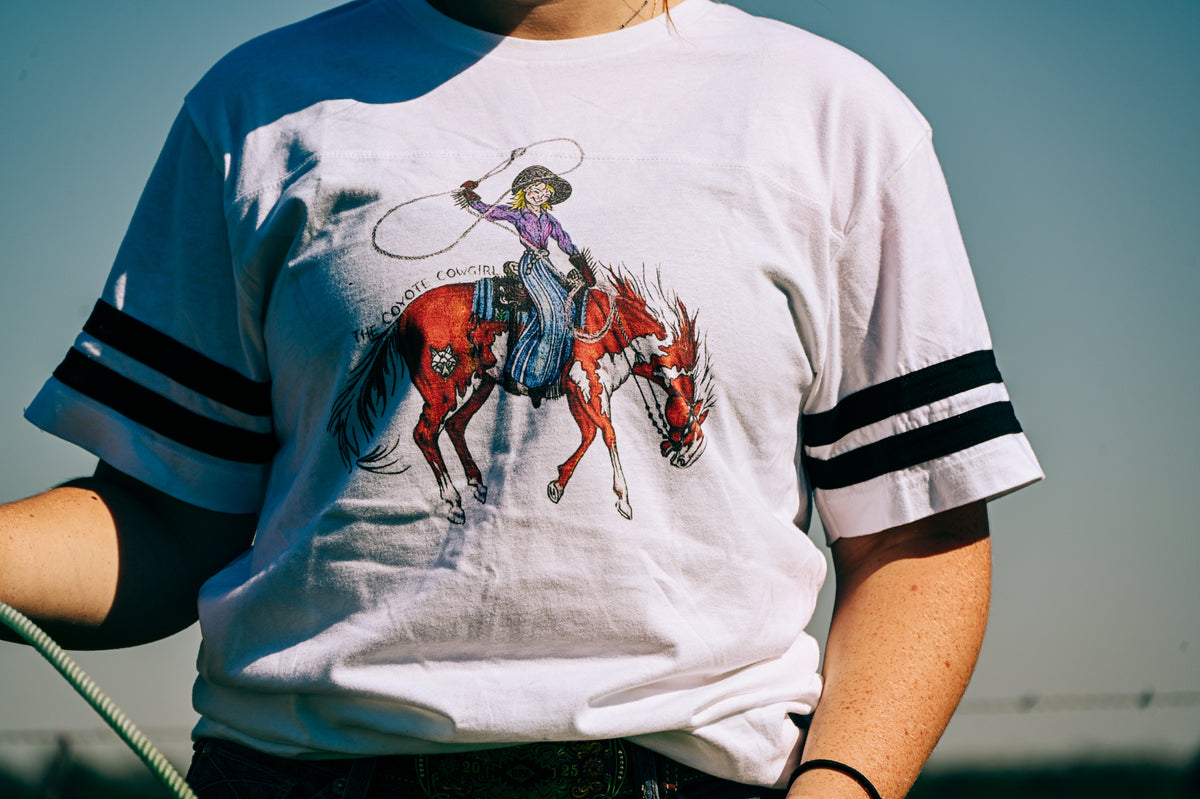 A person wearing a white short-sleeve t-shirt with black stripes on the sleeves and a graphic of a cowgirl riding a red horse on the front, photographed outdoors against a blue sky.