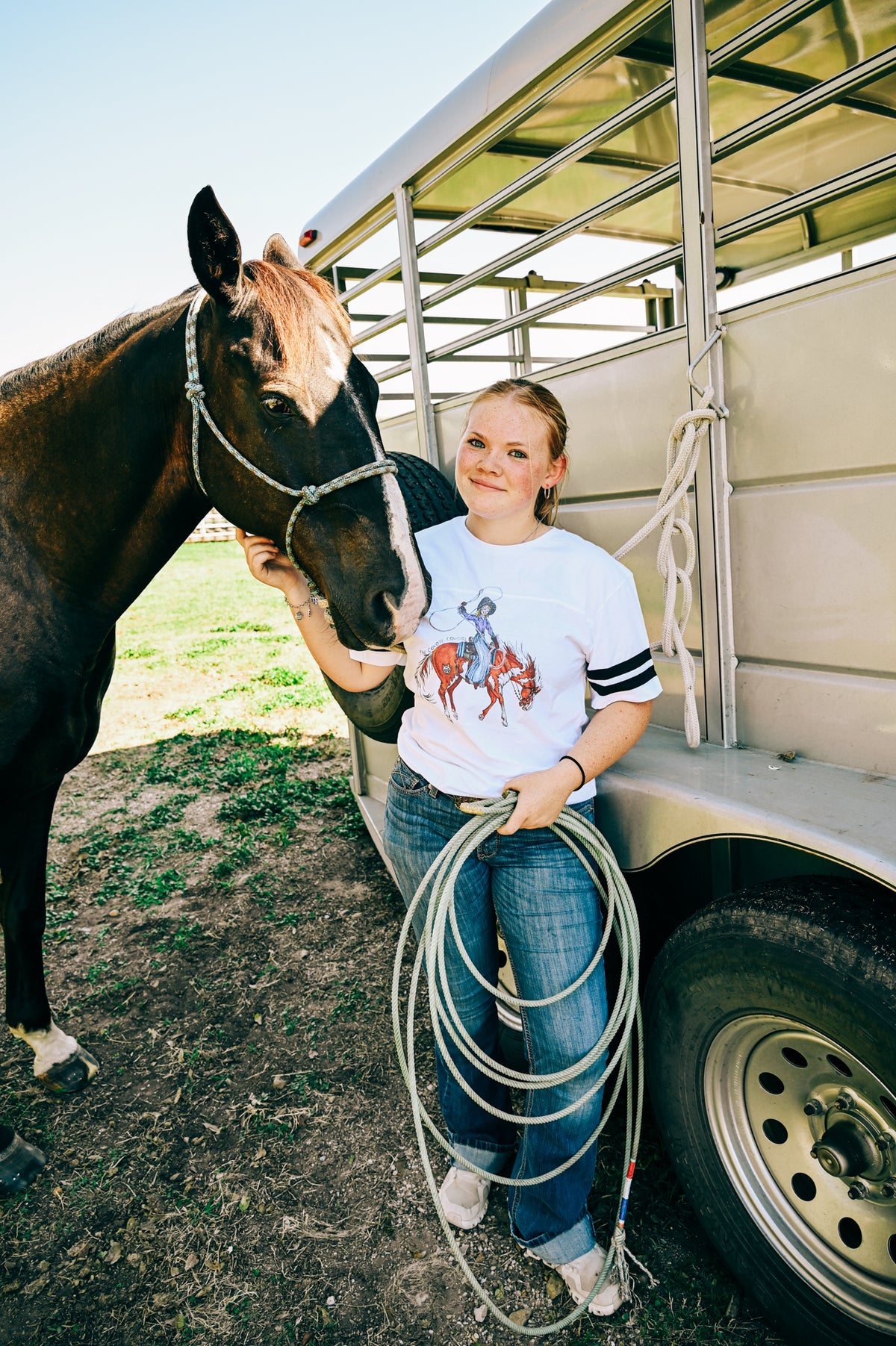 A young woman stands next to a horse and a trailer, wearing a white graphic t-shirt with a cowgirl riding design, blue jeans, and holding a coiled rope.