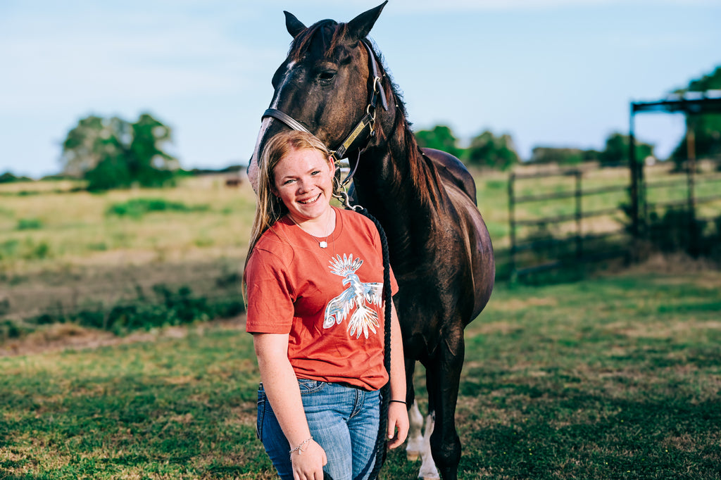 A smiling young woman in a Mauve Thunderbird graphic tee and blue jeans stands next to a dark brown horse in an outdoor grassy field under a blue sky.