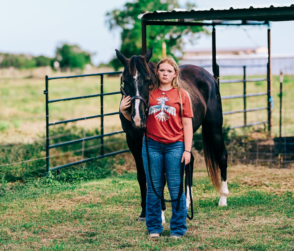 A young woman standing outdoors in a grassy field next to a dark brown horse, wearing a Women's  Mauve Western Thunderbird graphic tee and blue jeans.
