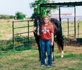 A young woman standing outdoors in a grassy field next to a dark brown horse, wearing a Women's  Mauve Western Thunderbird graphic tee and blue jeans.