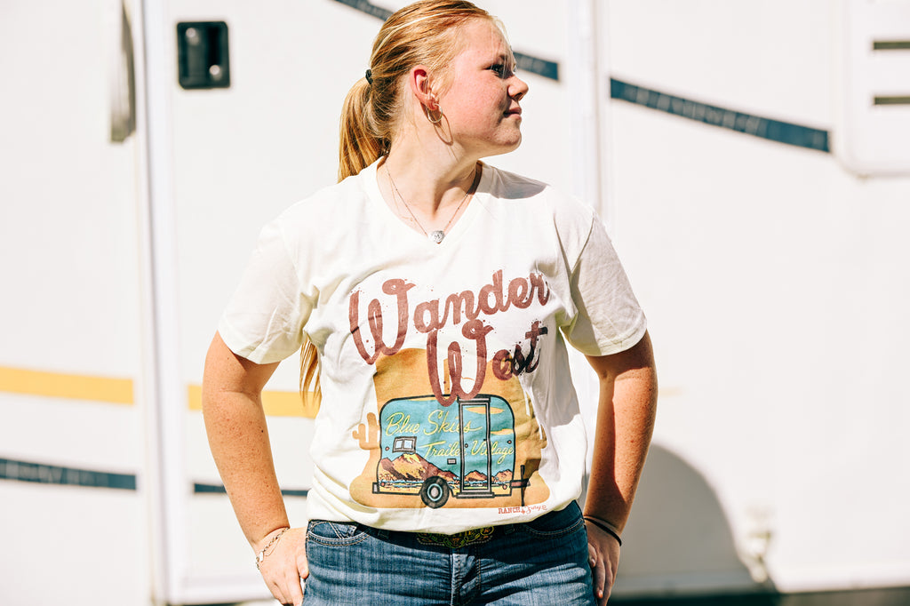 A woman wearing a cream-colored graphic t-shirt with 'Wander West' and a vintage RV design, paired with blue jeans, standing outdoors in front of a white RV.