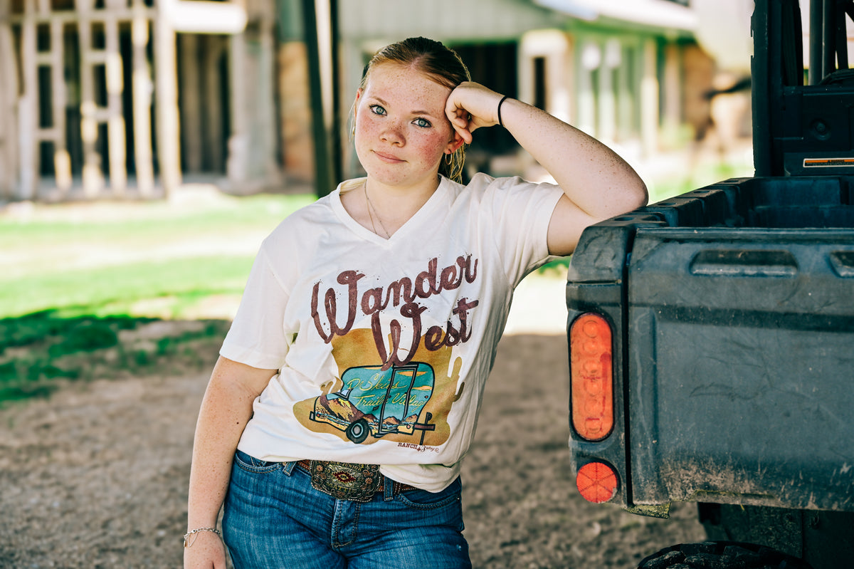 A woman wearing a cream-colored graphic t-shirt with 'Wander West' and a vintage RV design, paired with blue jeans leaning against a UTV bed.