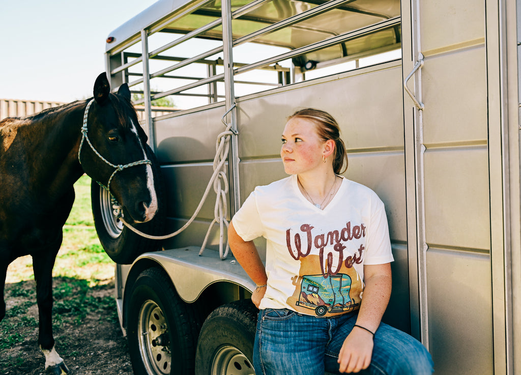 A young woman wearing a white graphic t-shirt that says 'Wander West' with a vintage camper illustration, sitting next to a horse trailer with a black horse beside her.