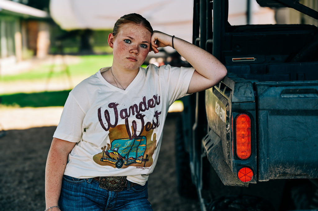 A young woman with freckles leans against the back of a dark pickup truck outdoors, wearing a white graphic t-shirt that reads 'Wander West' with a retro van illustration, paired with blue jeans.