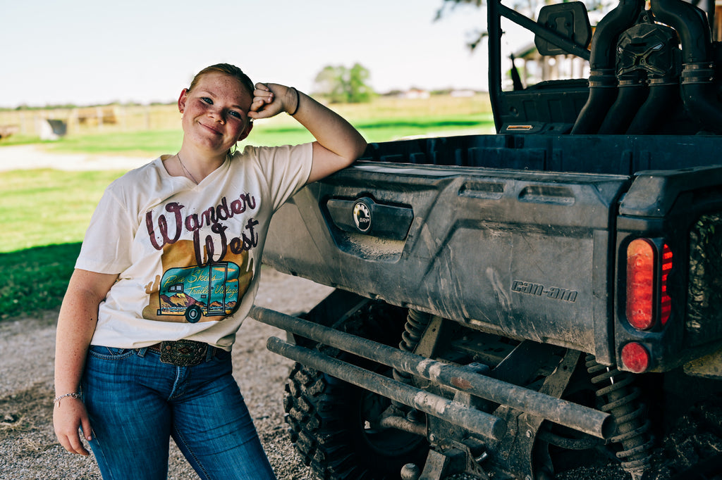 A young woman with freckles leans against the back of a dark pickup truck outdoors, wearing a white graphic t-shirt that reads 'Wander West' with a retro van illustration, paired with blue jeans.