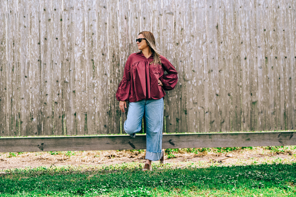 A woman stands outdoors against a weathered wooden fence, wearing a shiny burgundy long-sleeve blouse, light blue wide-leg jeans, and brown sandals.