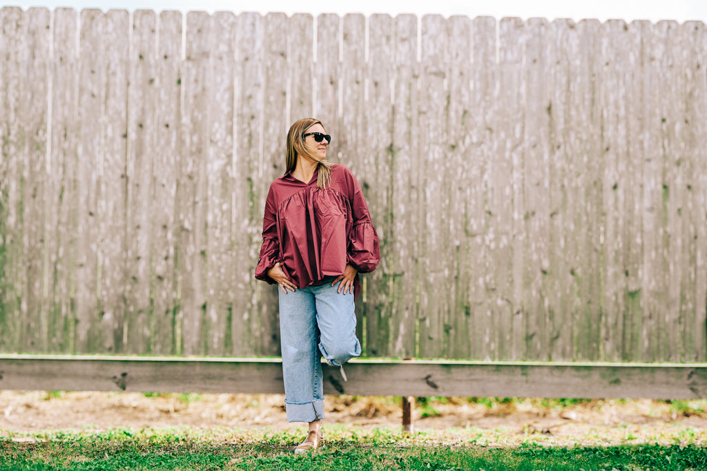 A woman wearing a burgundy long-sleeve blouse and light blue jeans stands against a weathered wooden fence outdoors.