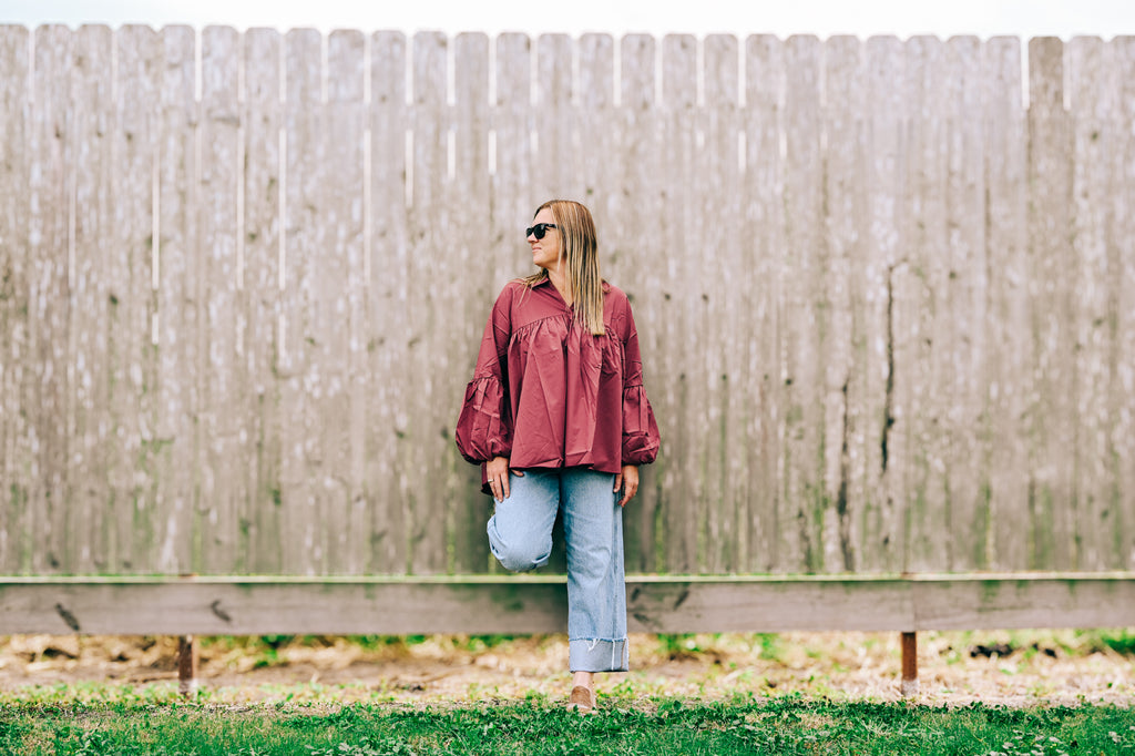 A woman wearing a wine flowy blouse, light blue jeans, and sunglasses stands against a tall weathered wooden fence.