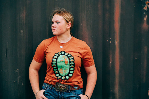A woman wearing an orange crewneck t-shirt with a large green and orange Southwestern-style geometric graphic on the front, paired with blue jeans and a western belt, standing against a dark corrugated metal wall.