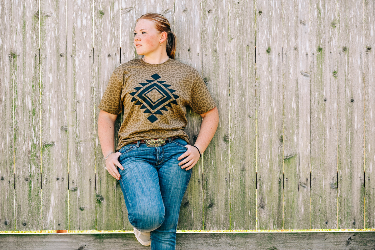 A young girl with blonde hair tied back stands against a weathered wooden fence, wearing a brown and black patterned t-shirt with a geometric diamond design, paired with blue jeans.