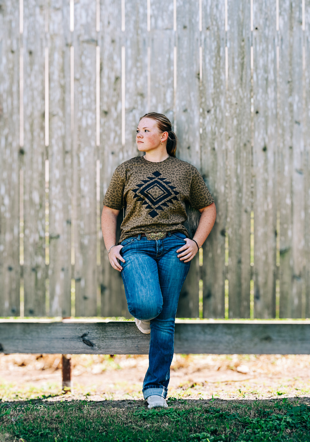 A woman stands outdoors against a weathered wooden fence, wearing a brown short-sleeve t-shirt with a black geometric diamond pattern, blue jeans, and white sneakers.