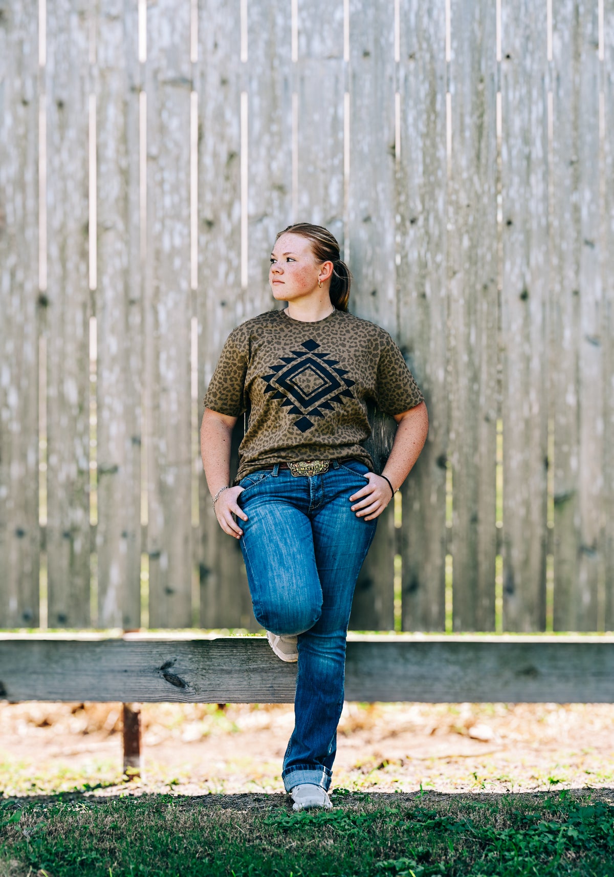 A woman stands outdoors against a weathered wooden fence, wearing a brown short-sleeve t-shirt with a black geometric diamond pattern, blue jeans, and white sneakers.