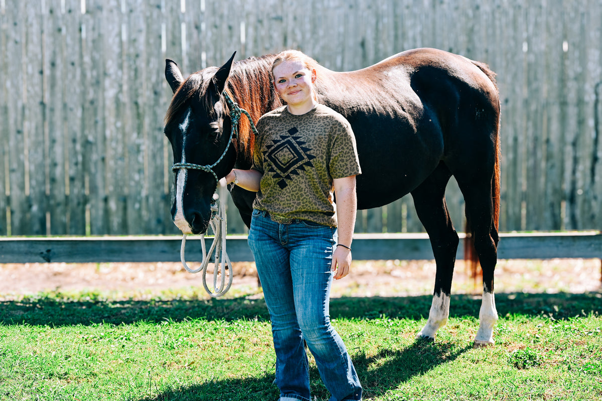 A woman standing outdoors on grass next to a dark brown horse, wearing a patterned olive-brown t-shirt and blue jeans, with a wooden fence in the background.