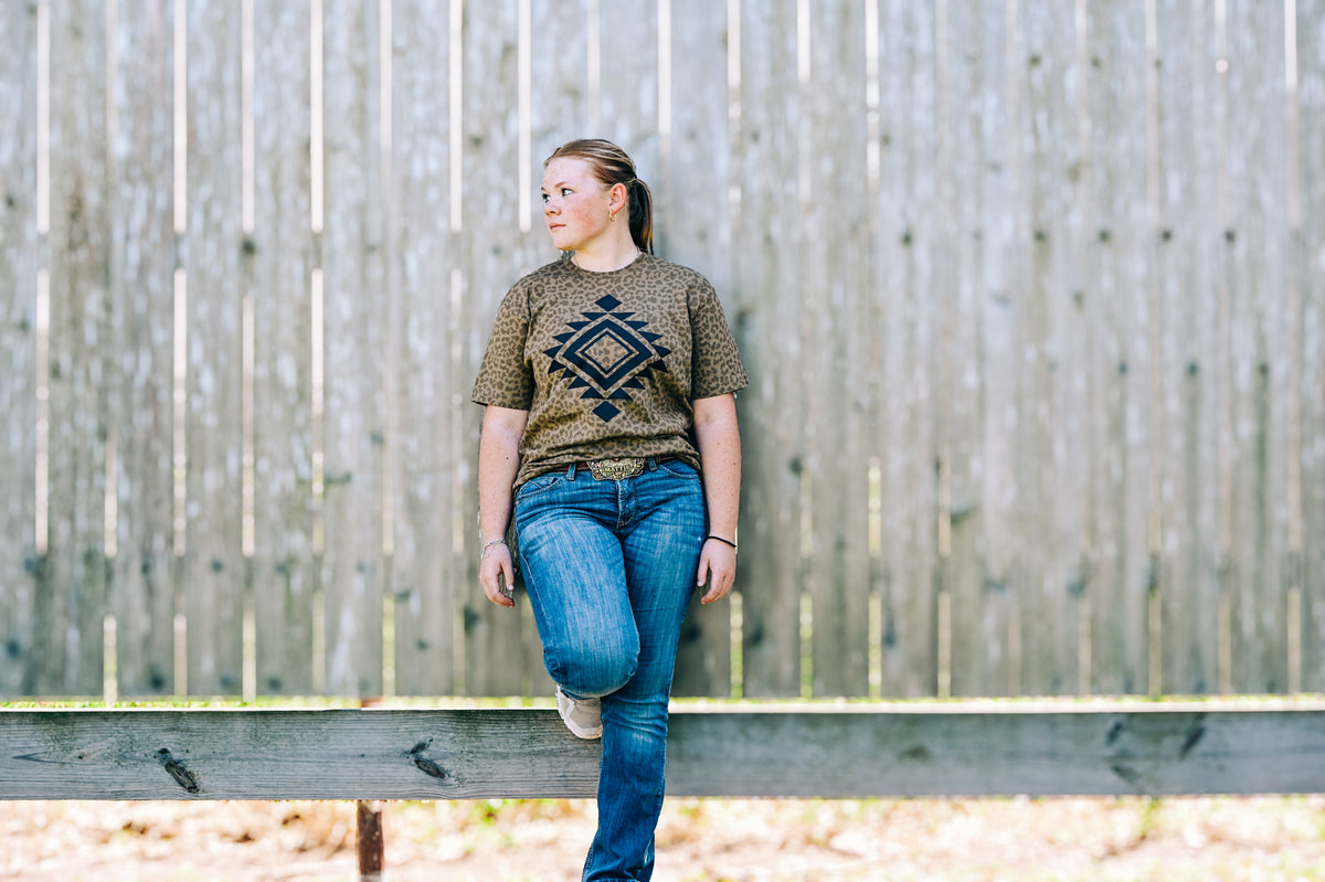 A woman wearing a brown patterned t-shirt with a black geometric diamond design and blue jeans, sitting on a wooden fence in front of a weathered wooden slat wall.