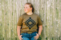A young girl with blonde hair tied back stands against a weathered wooden fence, wearing a brown and black patterned t-shirt with a geometric diamond design, paired with blue jeans.
