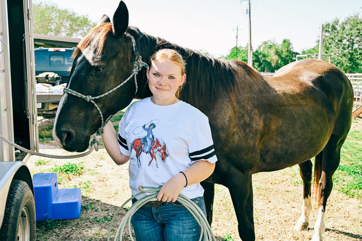 A young woman stands outdoors next to a dark brown horse, holding a coiled rope. She is wearing a white graphic t-shirt with black stripes on the sleeves and blue jeans.