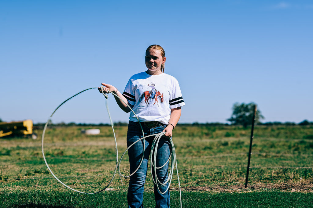 A woman stands in an open grassy field under a clear blue sky, holding a coiled lasso. She is wearing a white graphic t-shirt with black stripes on the sleeves and blue jeans.