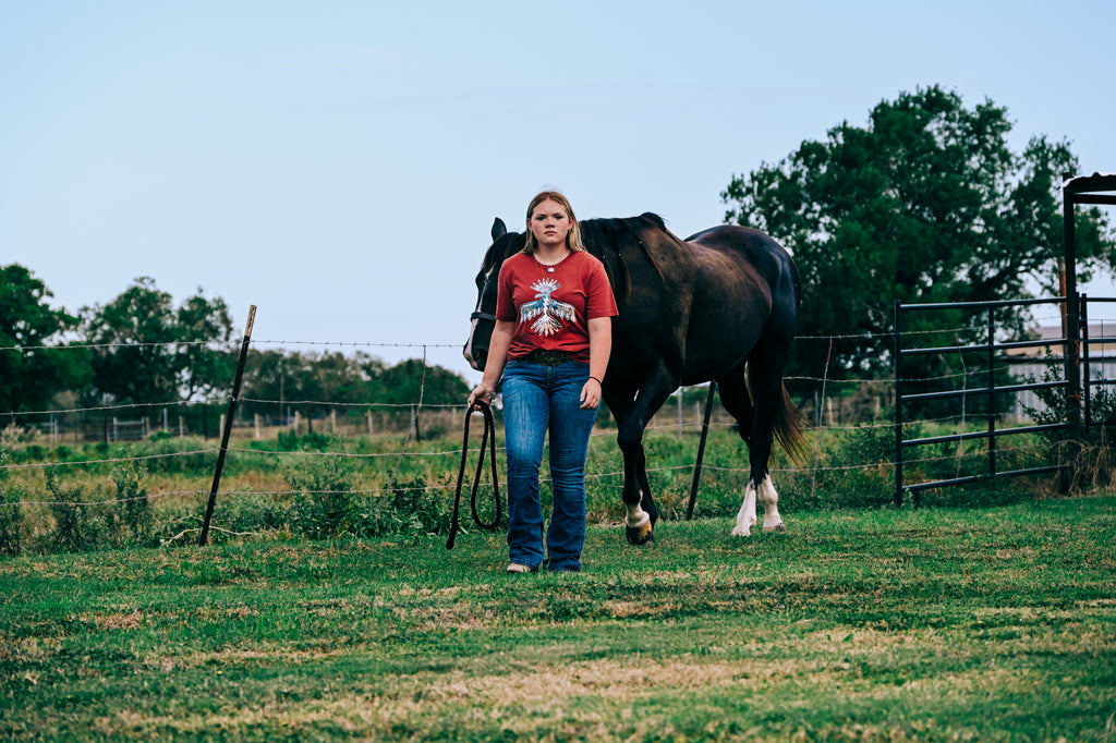 A young woman in a mauve Thunderbird graphic tee Western lifestyle outfit and blue jeans stands in a grassy field holding reins next to a dark brown horse, with trees and a fence in the background.