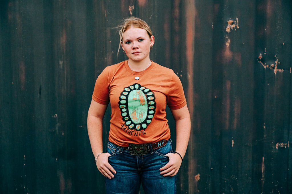 A young woman stands against a dark, weathered wooden wall wearing a rust-orange t-shirt with a large circular turquoise and green graphic on the front, paired with blue jeans.