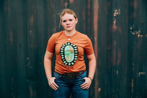 A young woman stands against a dark, weathered wooden wall wearing a rust-orange t-shirt with a large circular turquoise and green graphic on the front, paired with blue jeans.