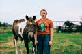 A woman standing in a grassy field next to a brown and white paint horse, wearing an orange graphic t-shirt with a turquoise ring that says authentic and blue jeans.
