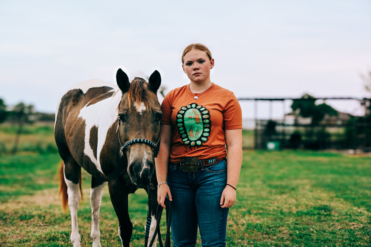 A woman standing in a grassy field next to a brown and white paint horse, wearing an orange graphic t-shirt with a turquoise ring that says authentic and blue jeans.