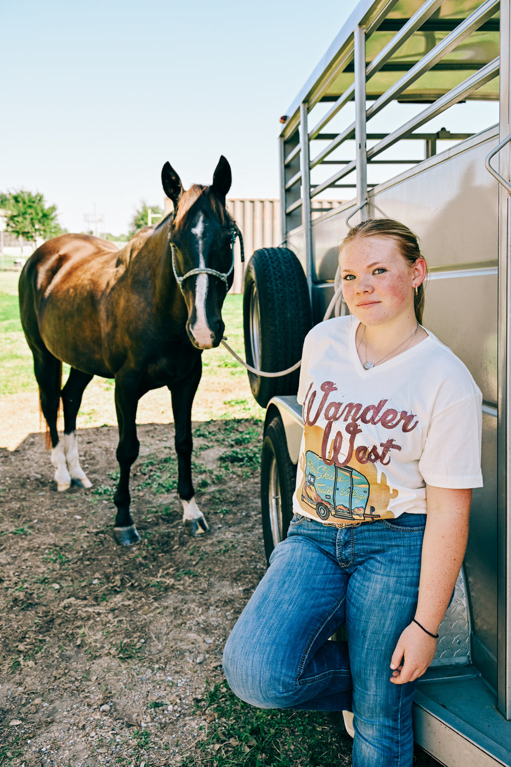 A young woman leans against a horse trailer next to a brown horse,wearing a cream-colored graphic t-shirt with 'Wander West' and a vintage RV design, paired with blue jeans.
