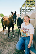 A young woman leans against a horse trailer next to a brown horse,wearing a cream-colored graphic t-shirt with 'Wander West' and a vintage RV design, paired with blue jeans.
