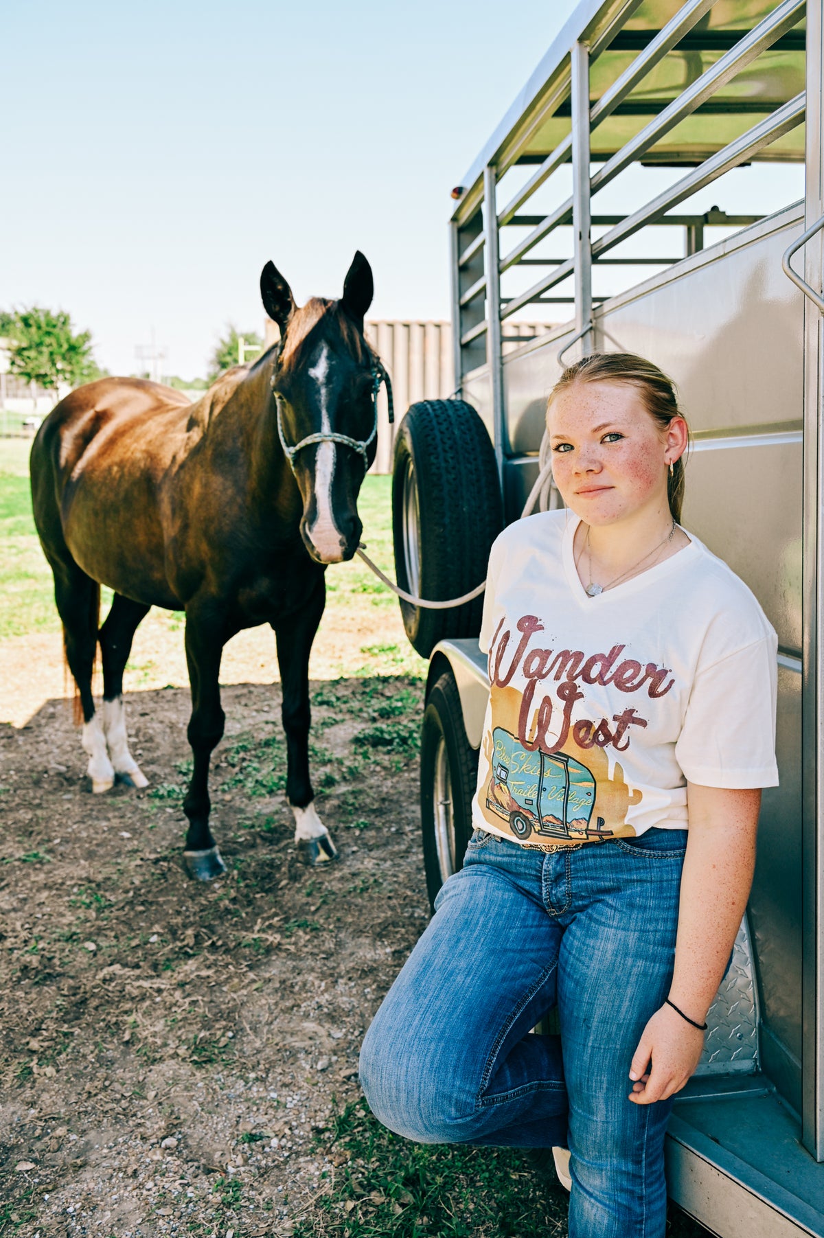A young woman leans against a horse trailer next to a brown horse,wearing a cream-colored graphic t-shirt with 'Wander West' and a vintage RV design, paired with blue jeans.