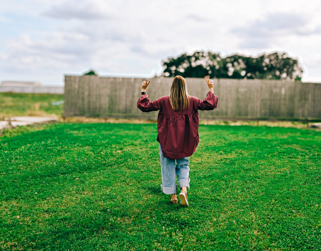 A woman seen from behind walking on a green grassy field with her arms raised, wearing a burgundy long-sleeve top, light blue jeans, and tan shoes, with a wooden fence and cloudy sky in the background.